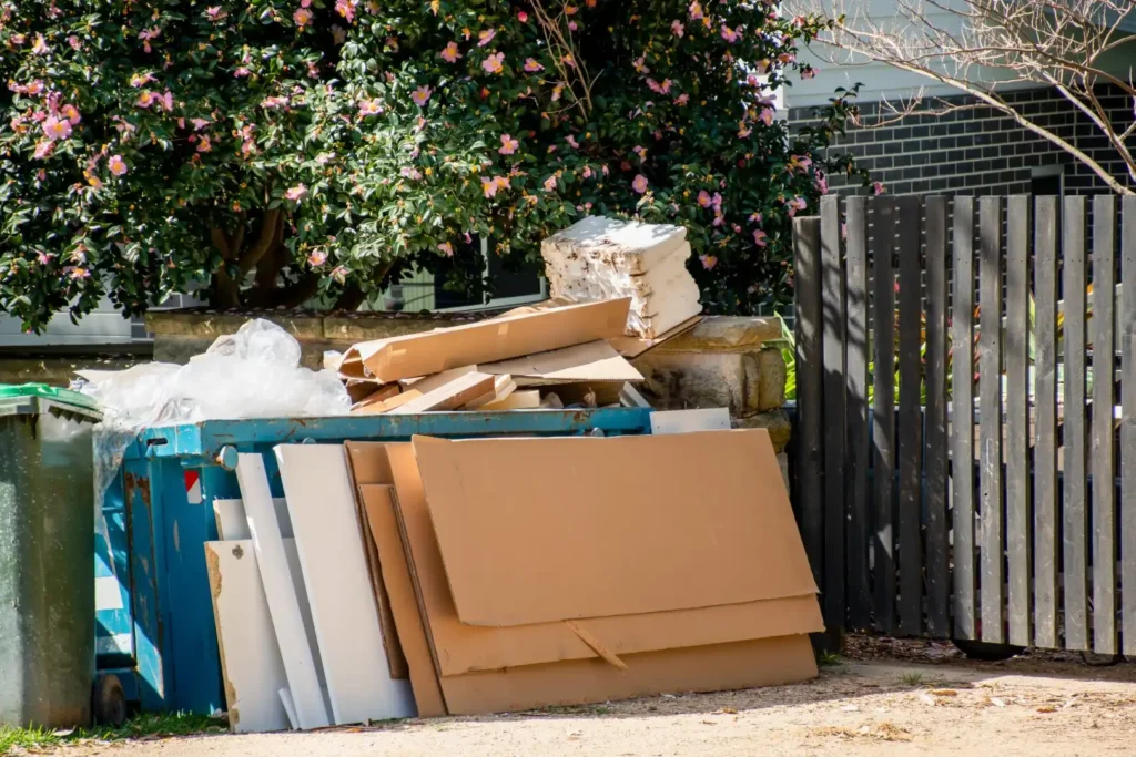 Discarded cardboard and packing materials are stacked next to a blue bin on a residential Winter Haven street. A flowering bush thrives near the grey wooden fence in the background, adding a touch of nature amidst the clutter.